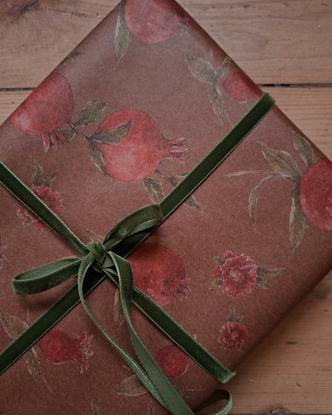 A gift box wrapped in brown kraft paper with a red pomegranate pattern and tied with a dark green ribbon, placed on a wooden surface.