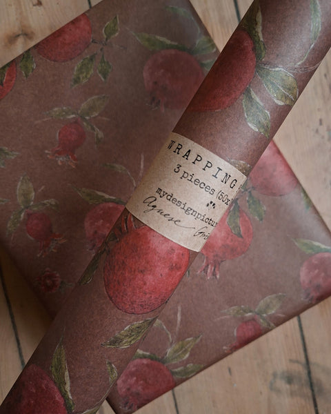 A roll of wrapping paper with a vintage pomegranate pattern on a brown background, wrapped around a gift box on a wooden surface. A paper label on the roll reads 'WRAPPING 3 pieces pack mydesignsbyAguene.