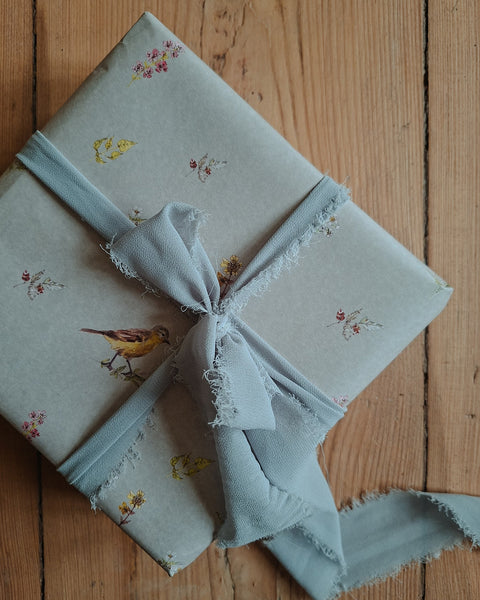 A gift box wrapped in light blue-gray fabric wrapping paper with a bird and floral print, tied with a matching frayed ribbon on a wooden surface.