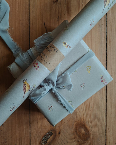 Light blue wrapping paper with a small bird and floral print, shown wrapped around gift boxes on a rustic wooden surface. One box is tied with a matching ribbon and bow, and a kraft paper band is visible.