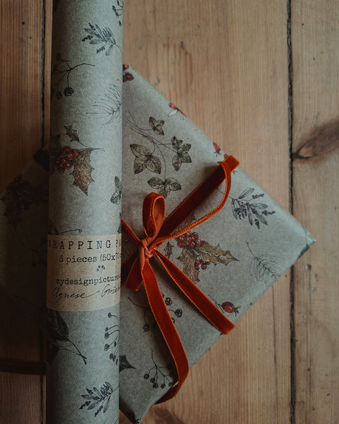 A roll of wrapping paper and a wrapped gift box on a rustic wooden surface. The wrapping paper has a muted sage green background with a vintage botanical pattern of holly leaves and red berries, and is tied with a bright orange velvet ribbon.